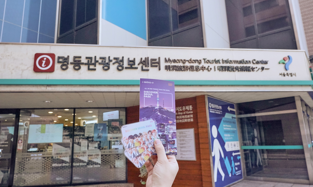 A photo of someone holding a Discover Seoul pass and a guidebook in front of the Myeong-dong Tourist Information Center.