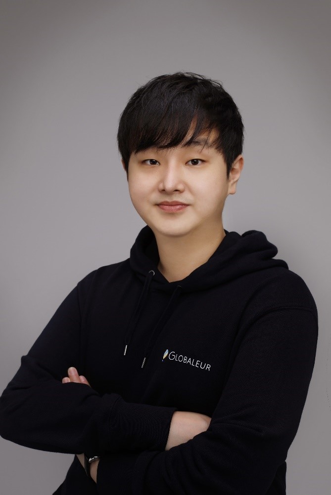 A man is posing in front of a gray background with his arms crossed, wearing a black hooded t-shirt.