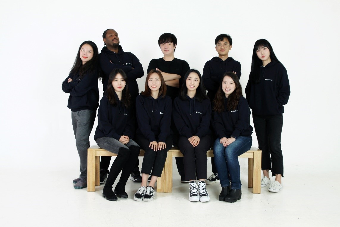 There are two rows of employees wearing black hooded t-shirts. One woman, three men, and one woman are standing in the back row from the left. In the front row are four women seated.