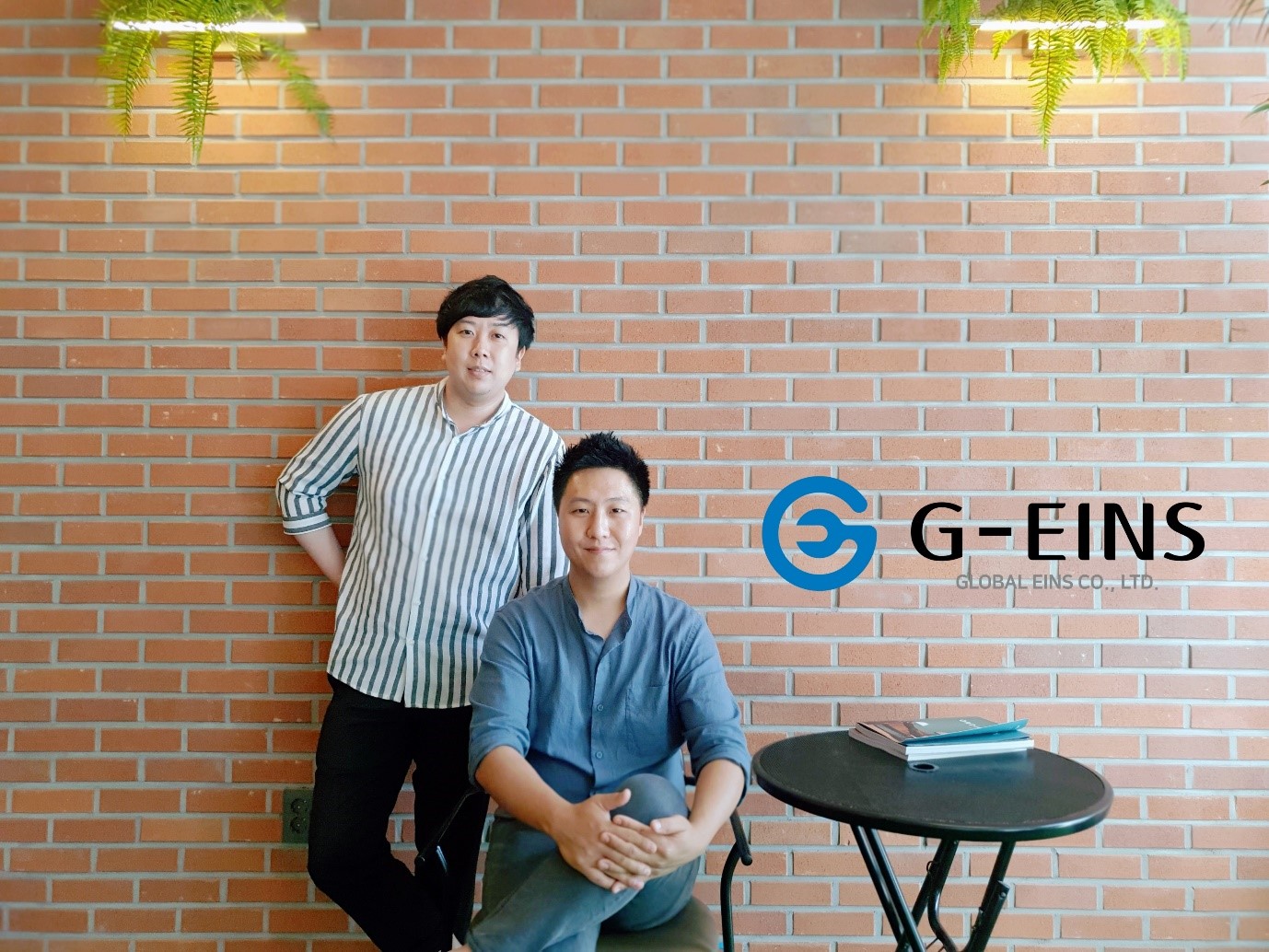 There are two men on the left side. The man on the left is standing, and the man on the right is seated. A black table can be seen to the right. There is a magazine on the table. On the table, the text “G-EINS” is written in black and “GLOBAL EINS CO., LTD.” in gray.