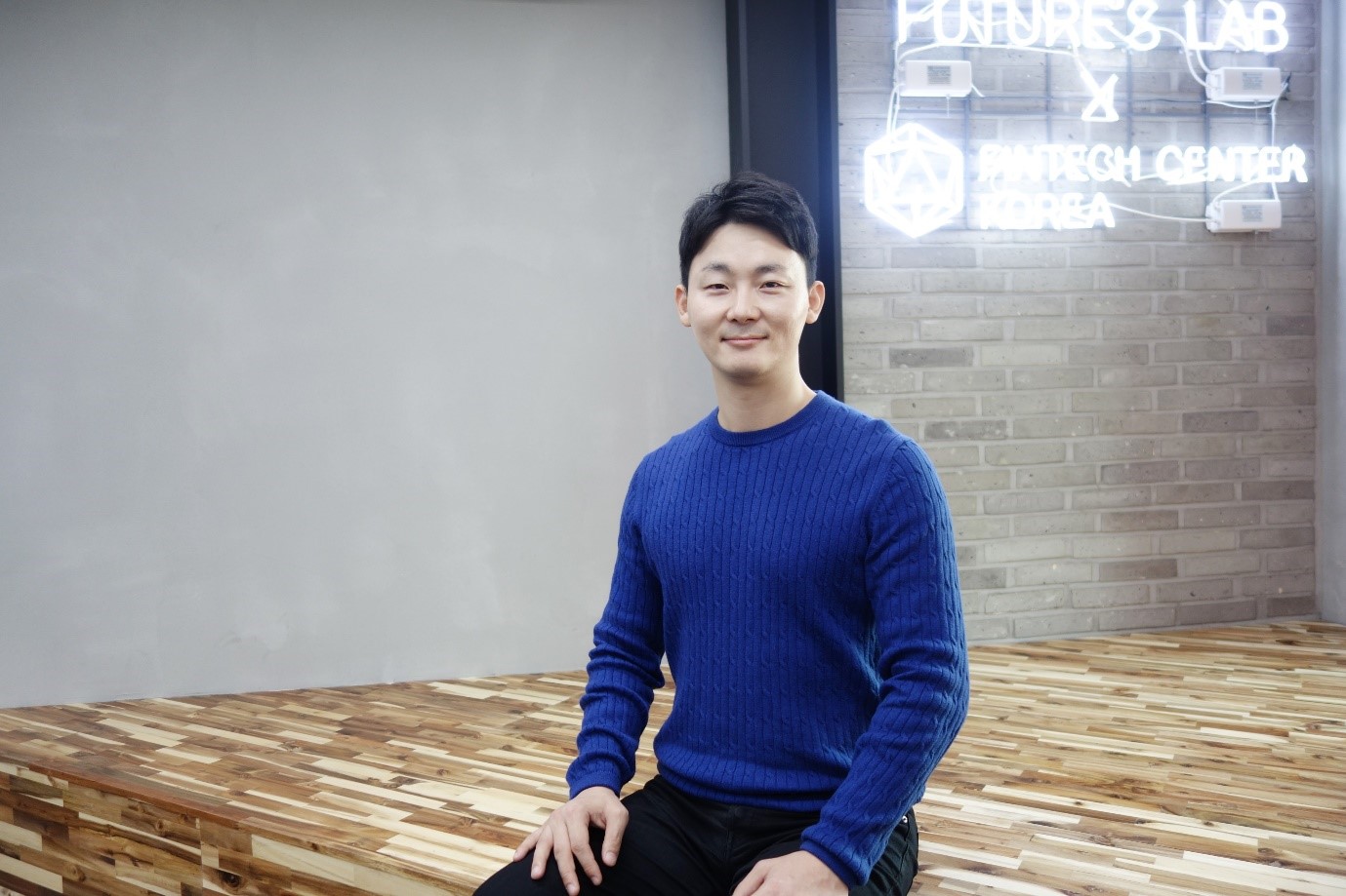 A man in a dark blue top and black bottoms is sitting on a wood-patterned floor.