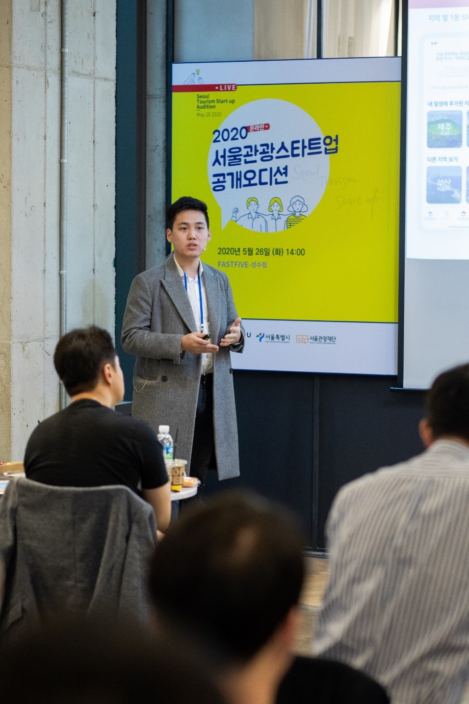 A man is giving a presentation using the microphone in front of a yellow and white poster with blue text reading “2020 Seoul Tourism Startup Audition, May 26, 2020 (Tuesday), 14:00.”In front of the man, three judges are listening to the presentation.