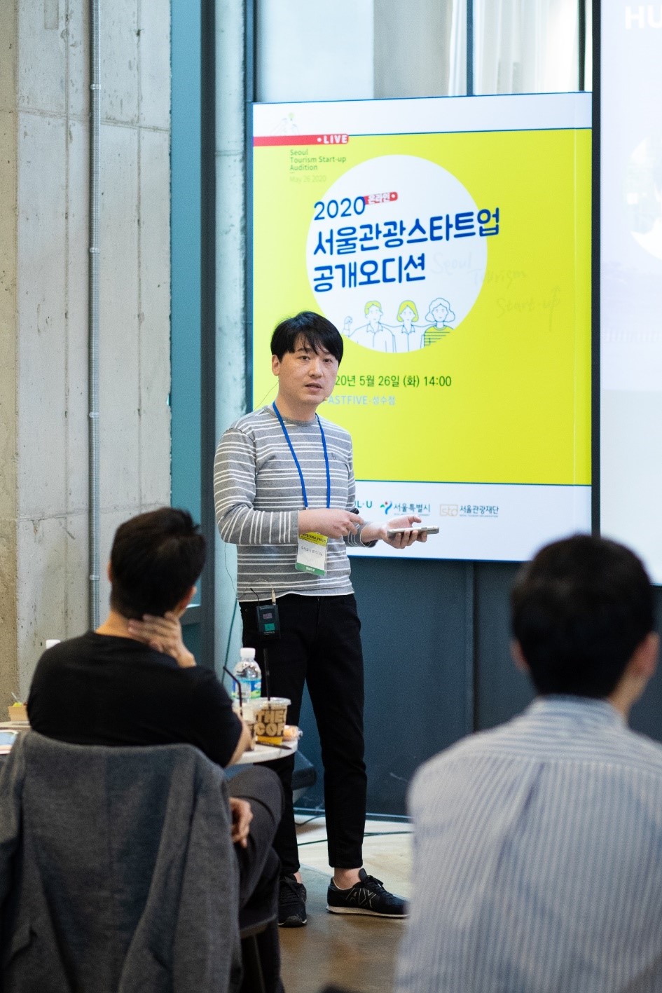 A man holding a wireless presentation pointer is giving a presentation using a microphone in front of a yellow and white poster with blue text reading “2020 Seoul Tourism Startup Audition, May 26, 2020 (Tuesday), 14:00.”