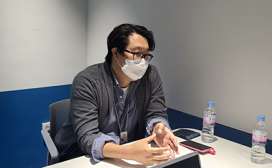 A man wearing a white mask and black glasses is sitting at a desk. On the desk are two bottles of mineral water, a dark blue handkerchief, and a smartphone in a pink case.