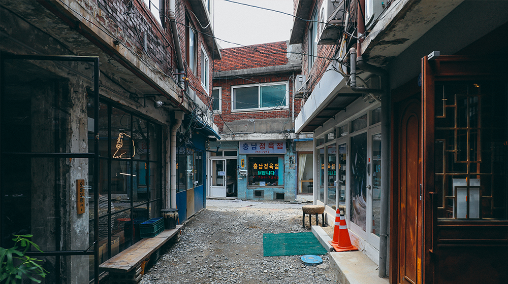 A dirt alleyway with stores and restaurants in operation on either side and a butchershop off in the distance named, Chungnam Jeongyukjeom