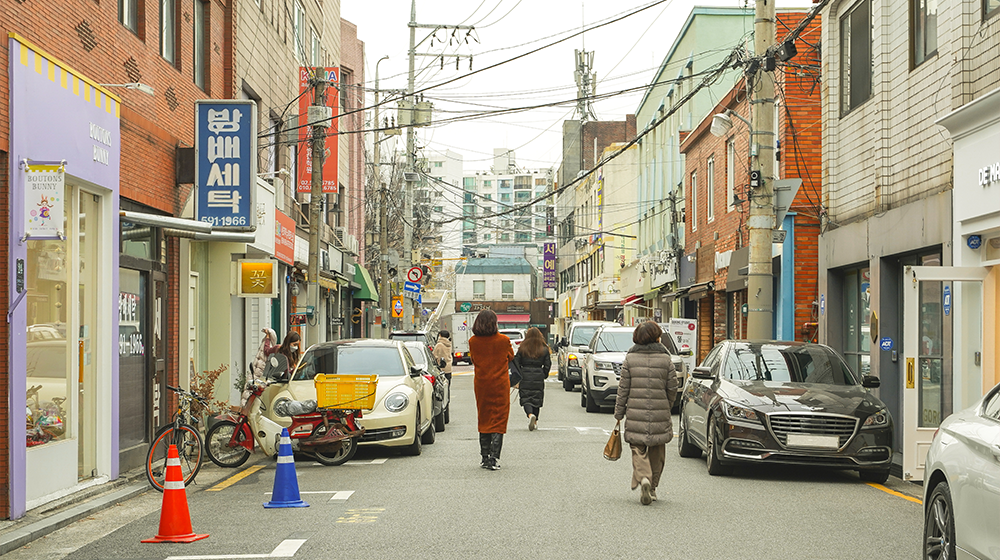 A sidestreet under several electrical wires and poles with people walking in the opposite direction and different stores and operations, including an accessory store, a flower store, and a laundrymat named, Bangbae Setak