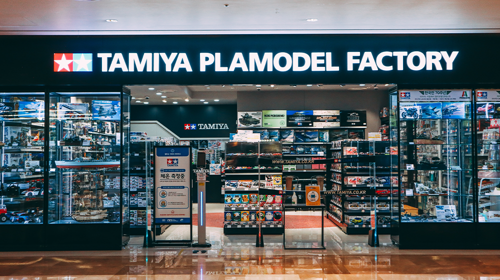 Tamiya Plamodel Factory store sign with the name in white lettering next to two stars on the left side and the entrance just below showing the various models on display