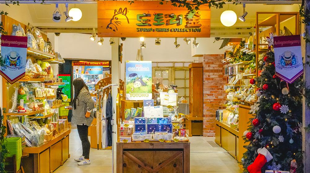 Studio Ghibli Collection written in green lettering on a wooden sign hanging in the middle of the store with all the various products and items on display across two separate aisles with a woman doing some shopping